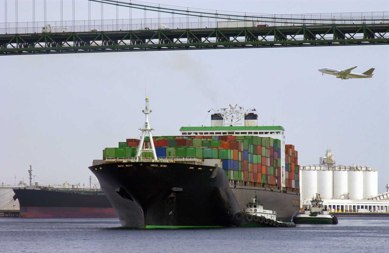 transport-port-of-long-beach-california-land-sea-air A container ship is escorted under a bridge by tugboat while trucks drive across a bridge and an aircraft flies overhead.