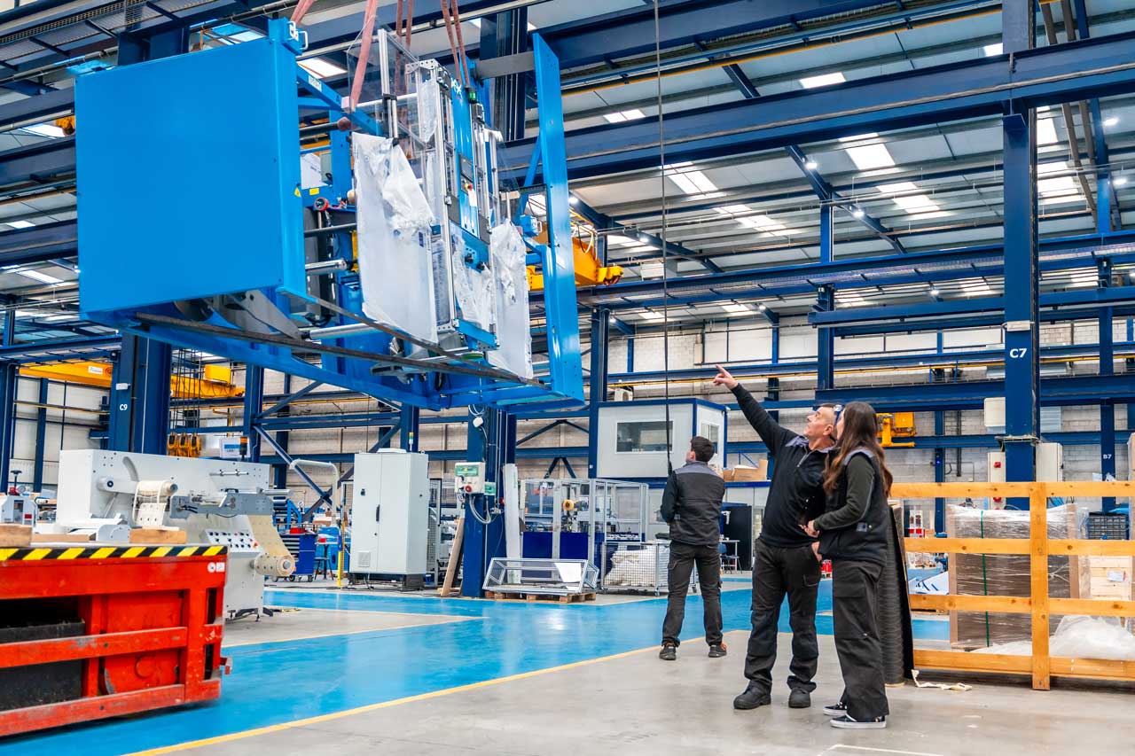 manufacturing-facility-lifted-metal-rack A man speaking with a woman points to a piece of machinery lifted in the air by a crane inside a manufacturing facility.